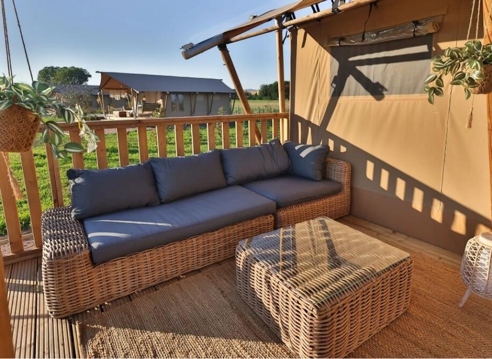 Veranda con divano in rattan, piante sospese e vista su tende glamping.