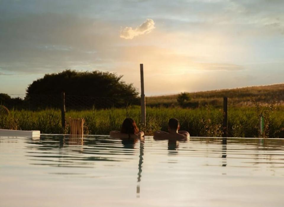 Due persone rilassate in piscina al tramonto con vista su un campo verde.